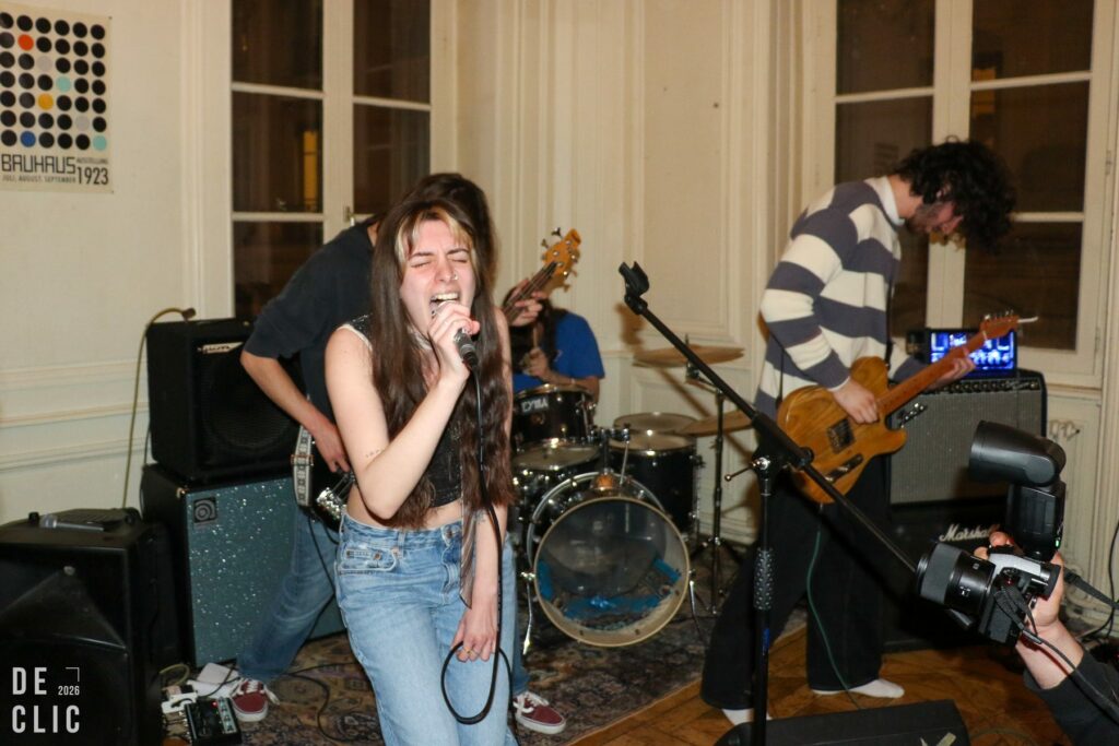 Une chanteuse au micro, très expressive, devant une batterie et un guitariste, dans le salon d'un appartement.