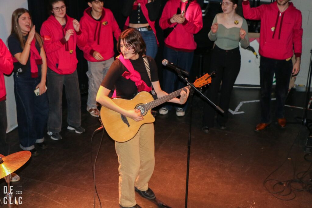 Une chanteuse s'accompagne à la guitare acoustique devant un micro sur pied, entourée de l'équipe en veste rouge.