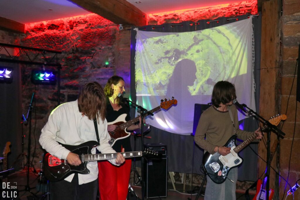 Trois musiciens à la guitare et à la basse jouent devant une projection vidéo verte, dans une salle aux murs de pierre éclairés en rouge.