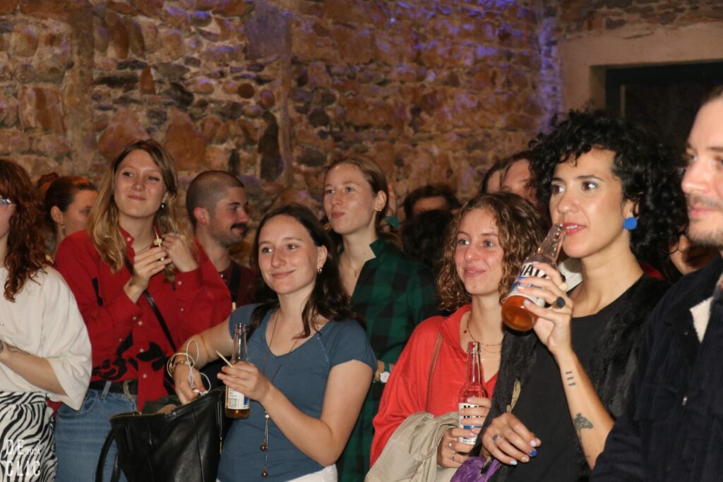 Le public, verre en main, écoute un concert dans une salle voûtée aux murs de pierre.