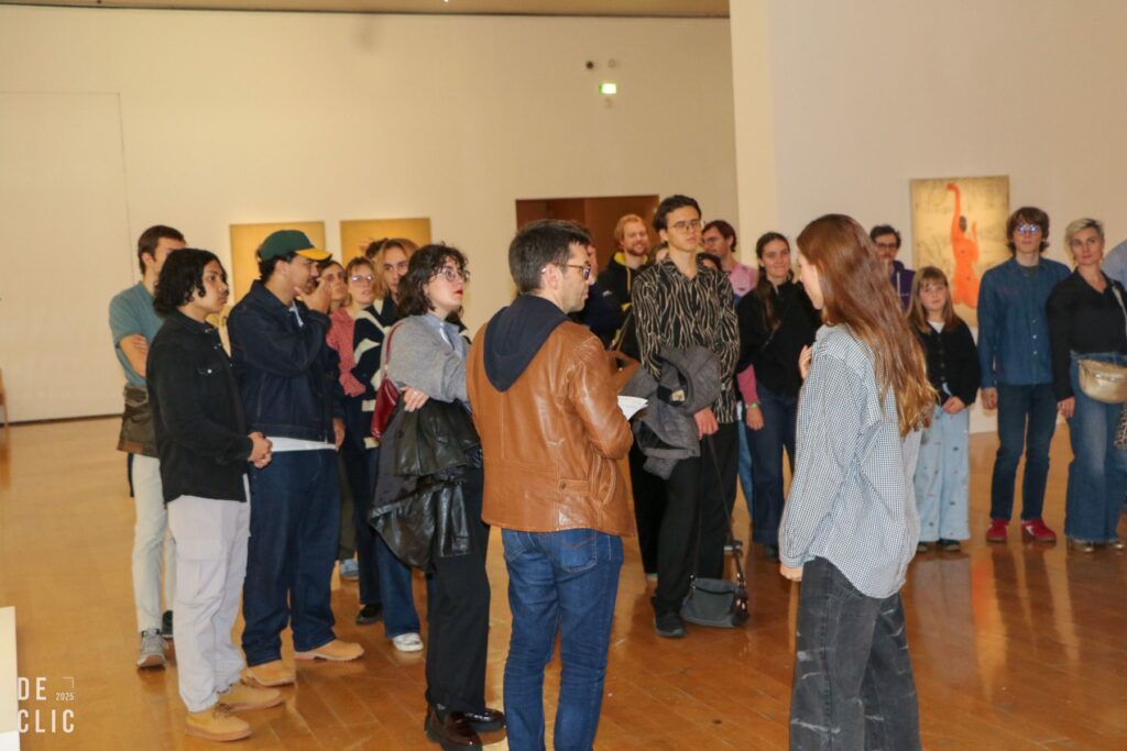 Une trentaine de visiteurs circulent dans une grande salle d'exposition au parquet clair, devant des tableaux colorés.